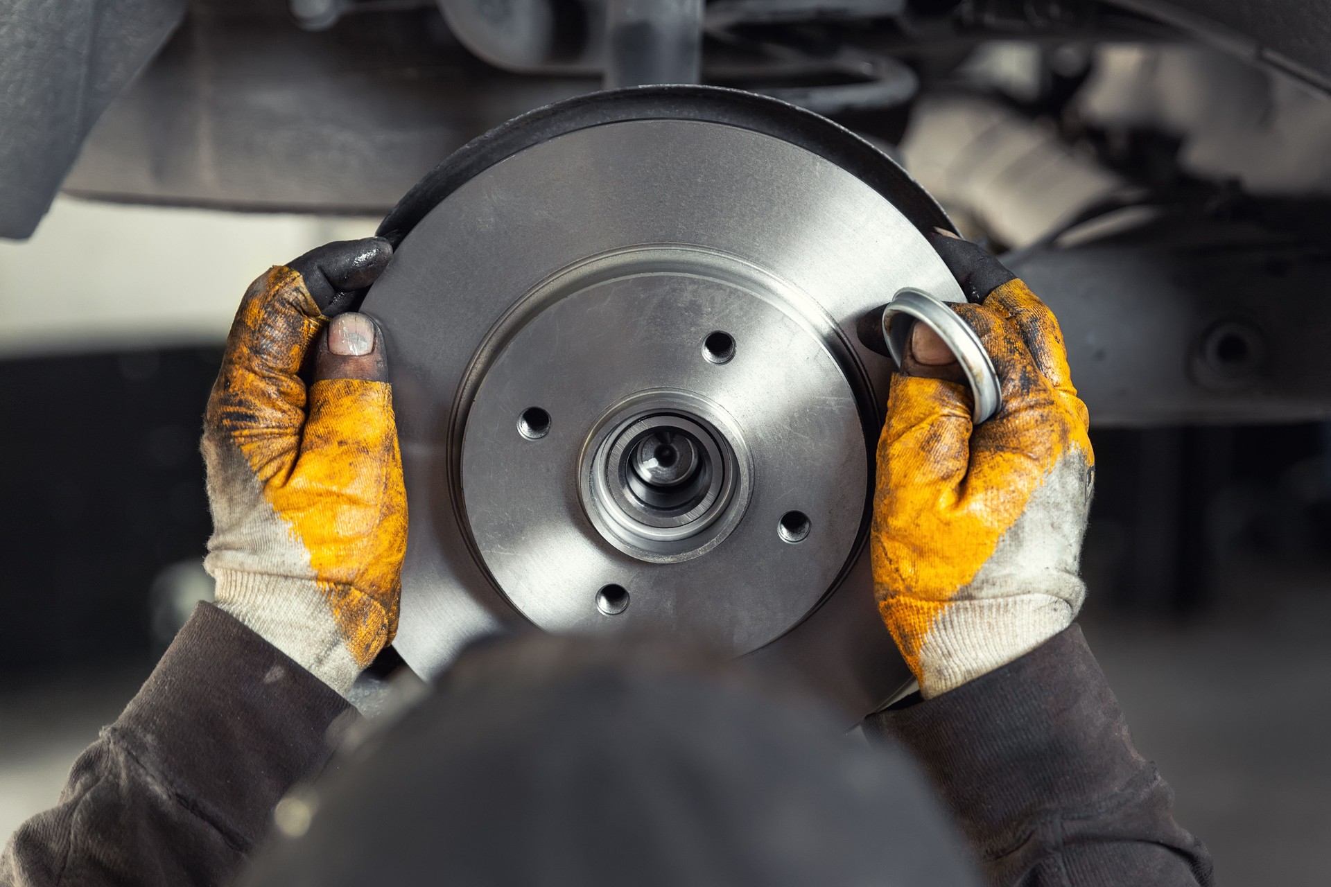 Closeup macho tehnician mecánico manos grasosas en guantes instalar nuevo disco de rotor de acero de freno oem coche durante el servicio en el centro de automóviles taller automotriz. Control de seguridad del vehículo y mantenimiento